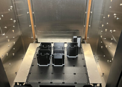 Blick in das Innere einer industriellen 3D-Druck-Anlage der Marke Stratasys. Auf der Druckplattform (Bauplattform) befinden sich mehrere schwarz gedruckte Kunststoffteile.