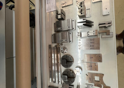 Eine Vielzahl unterschiedlicher, fertig bearbeiteter Metallbauteile liegt ordentlich sortiert auf einem weißen Arbeitstisch. Das Spektrum umfasst flache Aluminium- oder Stahlplatten mit Bohrungen, massive Blöcke, Leisten sowie runde Drehteile.