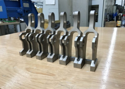 Sechs identische, komplex geformte Bauteile aus matt-grauem Metall stehen hintereinander auf einer hölzernen Werkbank.