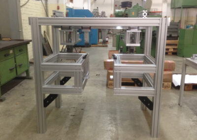 Ein mittig im Bild stehendes, teilweise montiertes Rahmen-Gestell aus silbernen Aluminium-Profilen in einer Werkstatt.