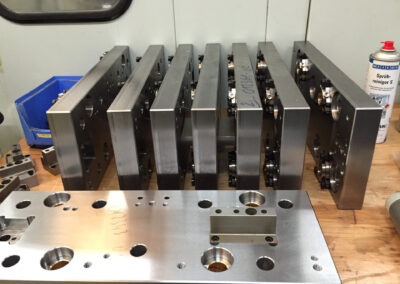 Auf einer hölzernen Werkbank stehen mehrere massive, rechteckige Stahlplatten hochkant hintereinander aufgereiht. Diese Platten sind Komponenten für Lochwerkzeuge und weisen präzise Bohrungen sowie montierte schwarze Befestigungselemente auf.