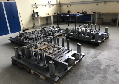 Drei massive, komplexe Transferwerkzeuge (Stanz- oder Umformwerkzeuge) aus Stahl stehen auf hölzernen Europaletten auf dem Betonboden einer hellen Werkstatt. Die Werkzeuge sind geprägt durch zahlreiche vertikale Führungssäulen, massive Grundplatten und teilweise goldfarbene Gleitelemente.