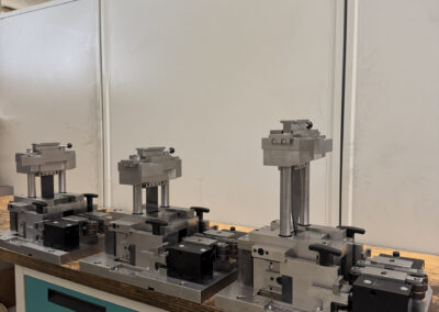 Drei identische oder sehr ähnliche, präzise gefertigte Trenn- oder Stanzwerkzeuge (möglicherweise Folgeverbundwerkzeuge) stehen auf einer hölzernen Werkbank mit türkisen Schubladen.