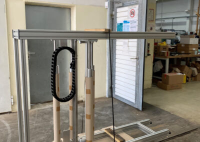 Eine unfertige oder demontierte Hebevorrichtung, konstruiert aus silbernen Aluminium-Konstruktionsprofilen, steht auf einer Holzpalette in einer Industrie- oder Lagerhalle.